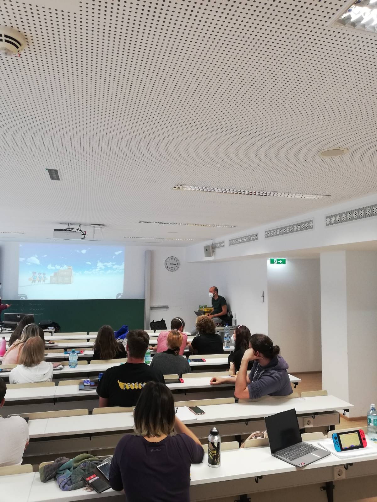 Foto eines Seminarraumes der Universität Wien. Studierende sitzen mit dem Rücken zur Kamera und beugen sich über Nintendo Switches. Auf die Projektionsleinwand ist ein von Studierenden erstelltes Level projiziert.