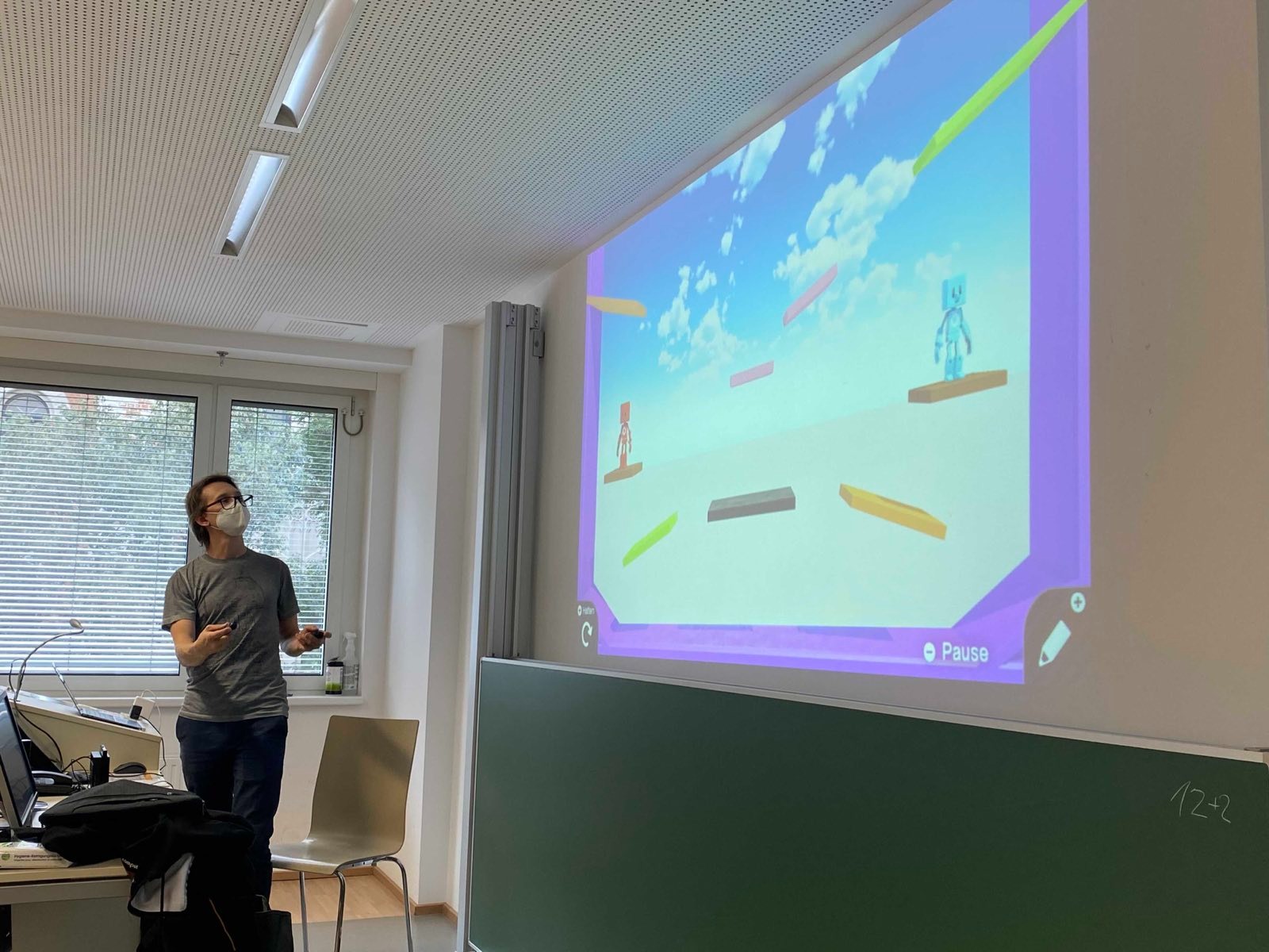 Foto einer Person vor der Projektionsleinwand eines Seminarraums der Universität Wien. Ein Mann hält zwei Nintendo-Switch-Controller in beiden Händen und gestikuliert vor der Projektionsleinwand. Projiziert wird ein Beispiel-Level.